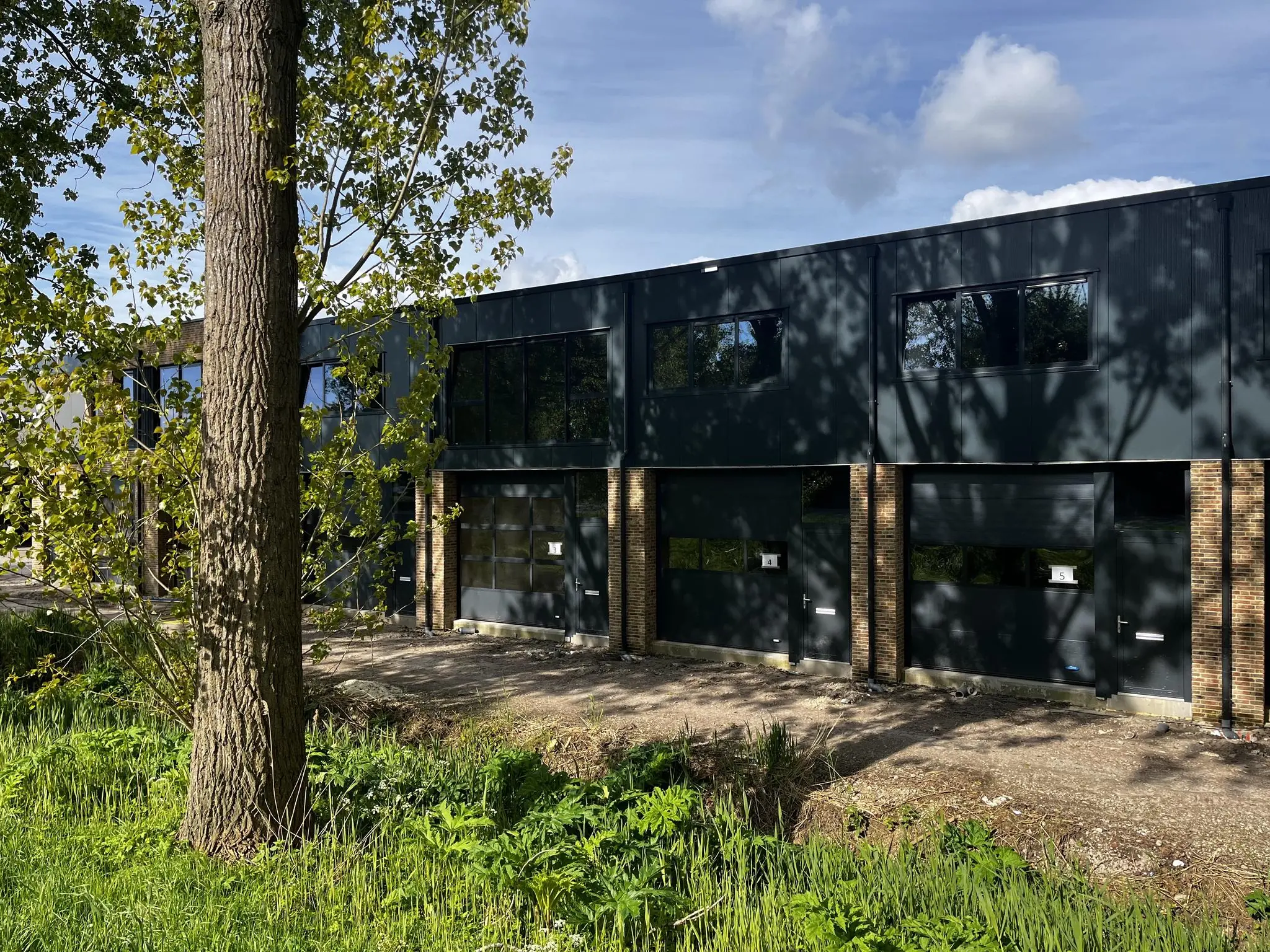 Bedrijfspanden aan Broekdijk West met donkere gevels en grote ramen, omgeven door groen en schaduw van bomen.